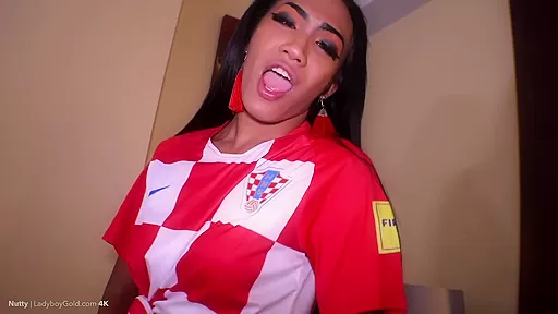 Ladyboy Nutty is wearing a white and red football jersey with knee high socks and gold heels